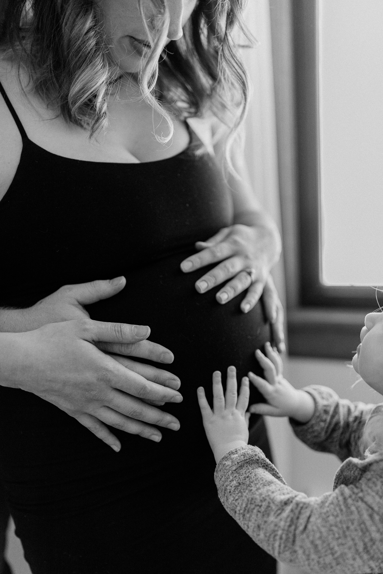Expectant mother holding her belly while her toddler reaches up to touch it, in soft black-and-white light